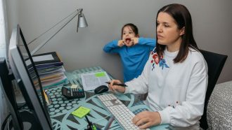 parent working on a computer while child makes a funny face in the background