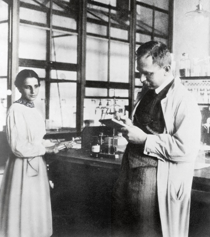 Lise Meitner stands next to a lab table and Otto Hahn holds a clipboard