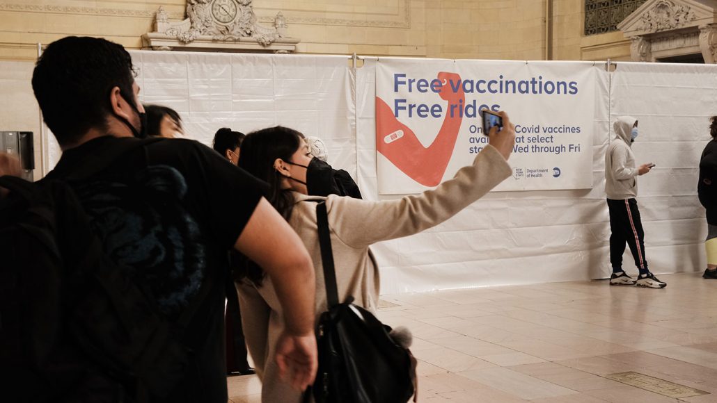 photo of six or so people in Grand Central Station wearing facemasks. a woman in the center takes a selfie