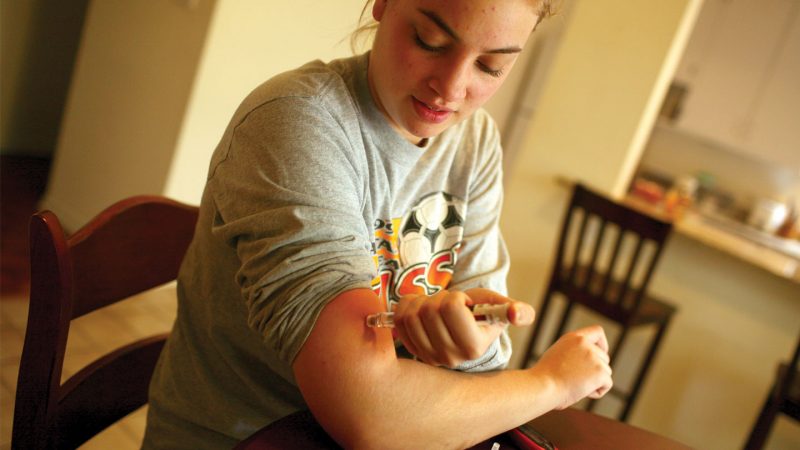 A young woman sitting at a kitchen table injects insulin into her arm