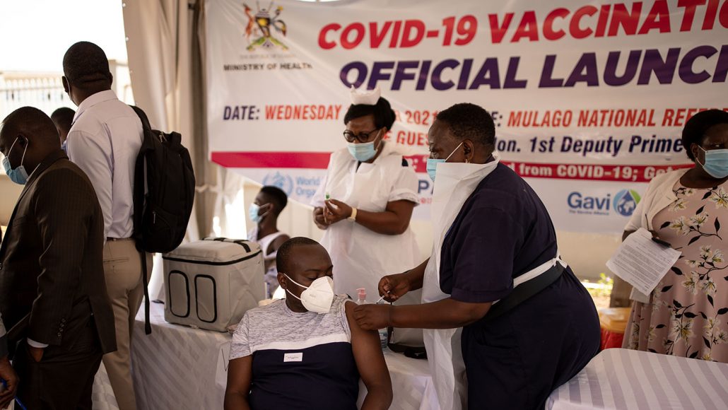 A woman in a mask and a disposable apron injects a vaccine into the arm of a man who is seated.