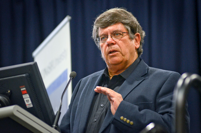 a man with dark hair and glasses wearing a sport coat speaking at a lectern