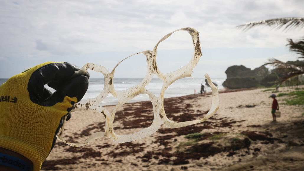 a gloved hand holds plastic pack rings (to hold six canned beverages together), behind the rings is a beach