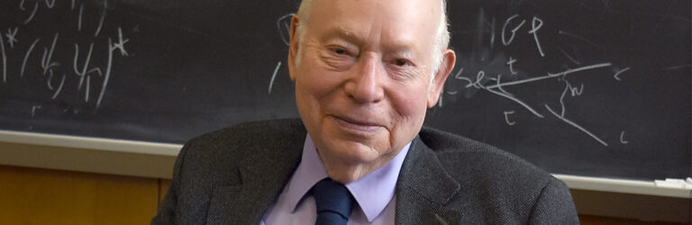 Steven Weinberg sitting in front of a chalkboard covered in equations