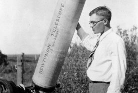 black and white photo of a young Clyde Tombaugh standing next to a telescope