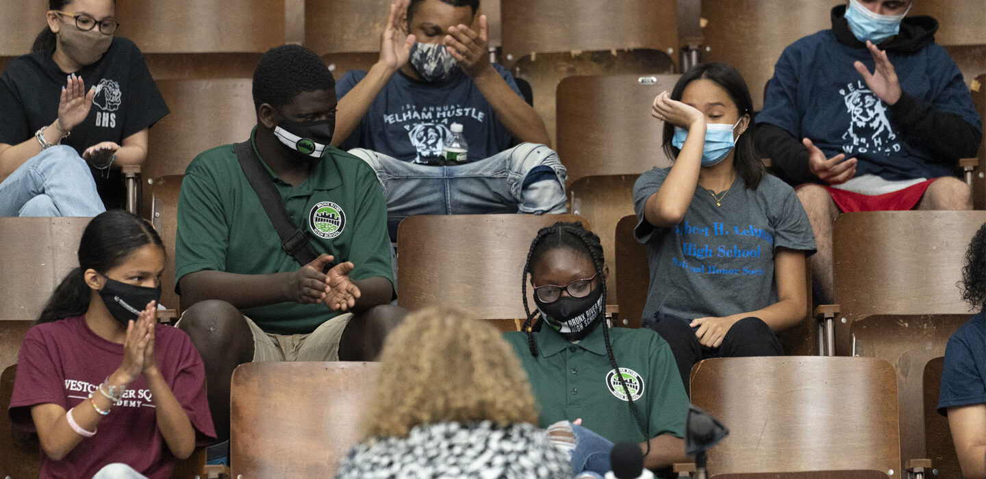 A small group of students in a lecture hall, wearing masks, with empty chairs between them. Several are applauding