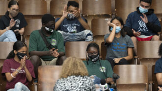 A small group of students in a lecture hall, wearing masks, with empty chairs between them. Several are applauding
