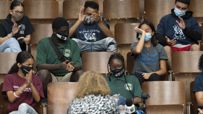 A small group of students in a lecture hall, wearing masks, with empty chairs between them. Several are applauding