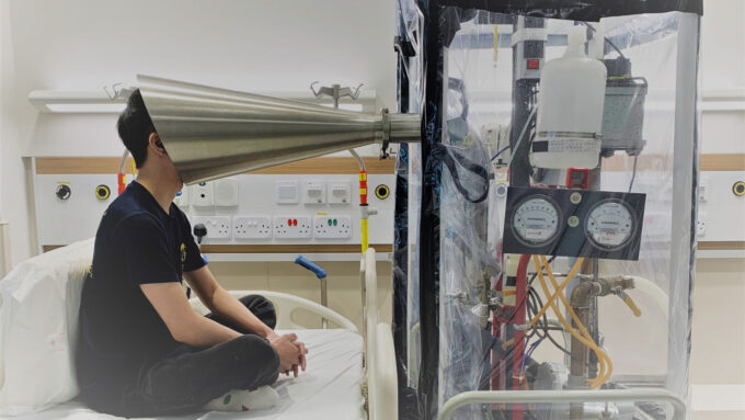 a researcher sits crosslegged on a hospital bed with their face inside a silver cone that's connected to a transparent chamber full of scientific apparatus