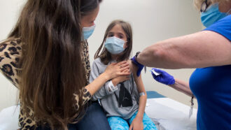 a small girl sits with her mom in an exam room as a nurse administers Pfizer's COVID-19 vaccine