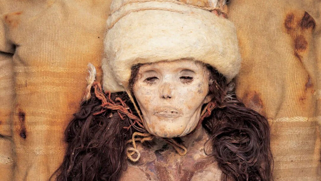 Het hoofd en de nek van een van nature gemummificeerde vrouw met lang donker haar, met een witte hoed op