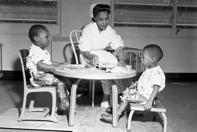 Black and white photo of a nurse sitting at a children's table with two small kids in leg braces