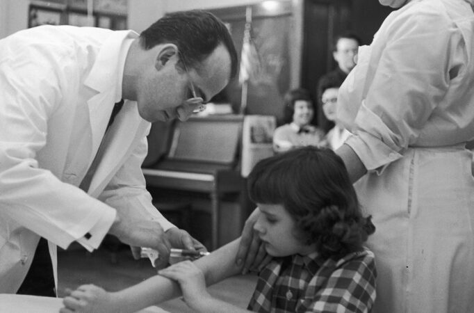 a man in glasses and a white coat gives a shot to a little girl