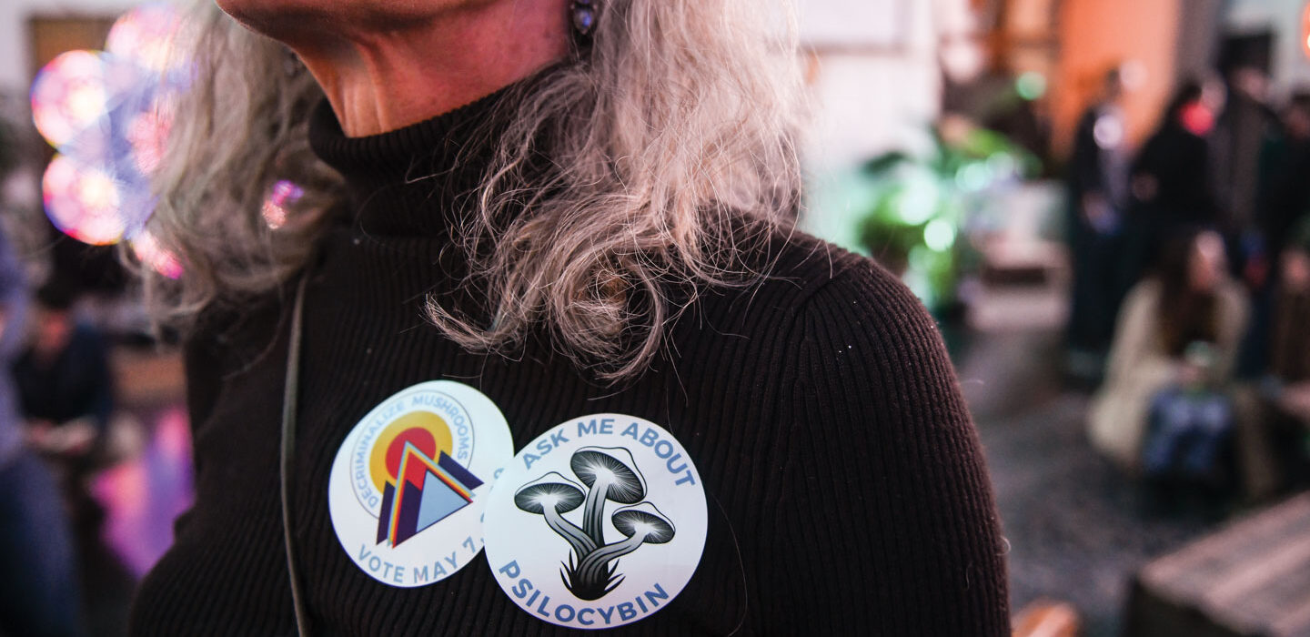image of a woman wearing a sticker that reads "Ask me about psilocybin" and another sticker that reads "Decriminalize Mushrooms"