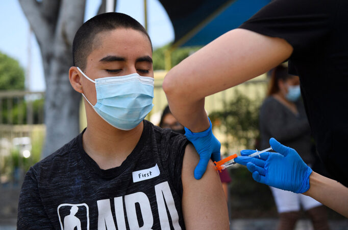 A teenager in a mask receives a vaccine shot from a nurse