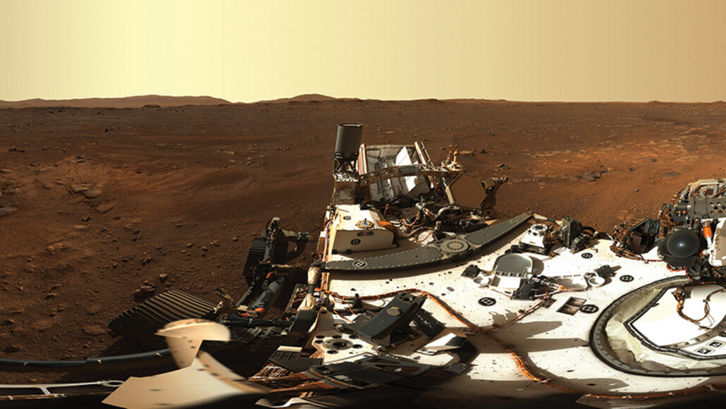 The rover Perseverance sits in the foreground with the red, rocky landscape and red-tinted sky in the background