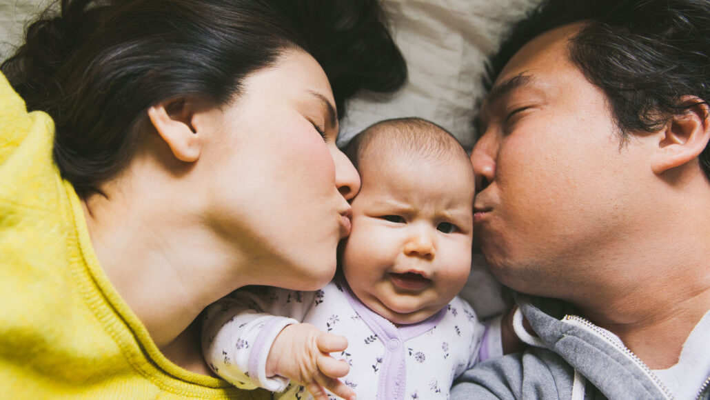 image of parents laying on either side of an infant and kissing the infant