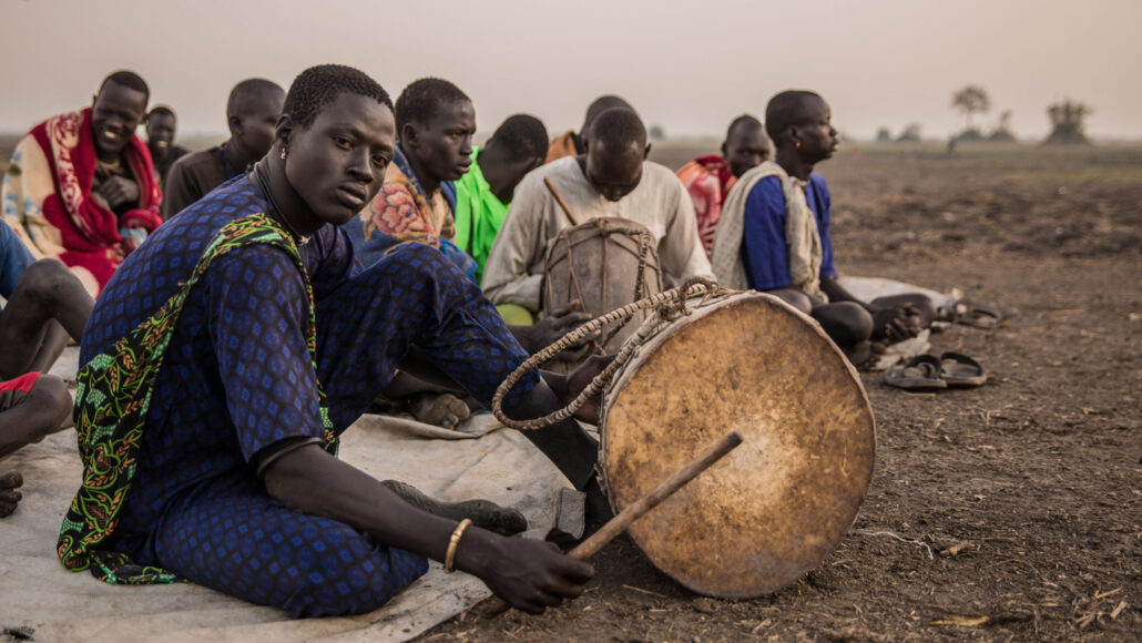 a group of Dinka people sitting on the ground where one man dressed in blue holds a drum and others hold instruments