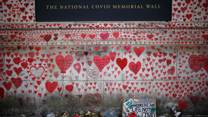 photo of a COVID-19 memorial wall painted with red hearts of varying sizes and messages; flowers and posters appear below