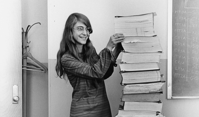 A black-and-white photo of Margaret Hamilton standing and smiling next to a very tall stack of books