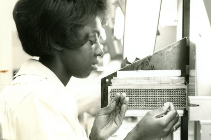an old, black-and-white photo of a woman threading metal wire through small holes in a machine