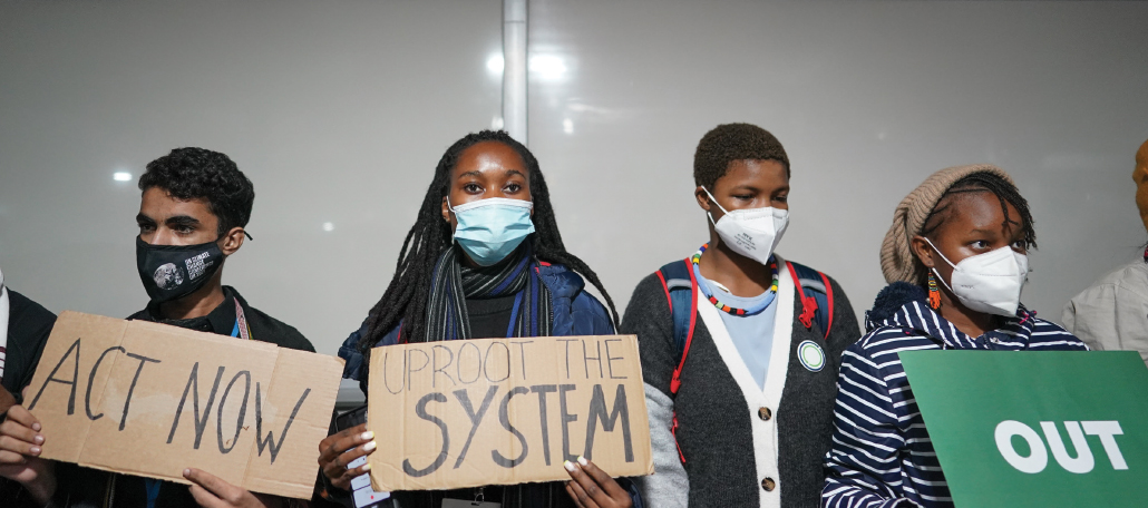 photo of young climate activists holding posters that read "Act Now" and "Uproot the system"
