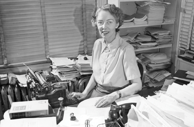 black and white image of Jane Stafford sitting at a desk with a typewriter and stacks of paper