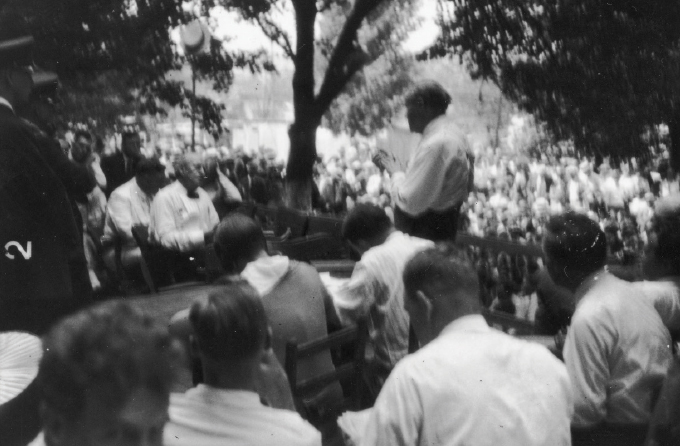 black and white image of a crowd seated around Clarence Darrow who is standing with his back to the camera and William Jennings Bryan who is seated