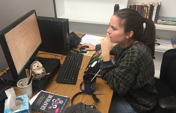 a photo of Maria Temming at her desk working. She is wearing a fuzzy plaid sweater, her hair is pulled back into a ponytail, and she is looking intently at her computer monitor. On her desk are a computer monitor, computer, phone, laptop, headphones, a book about graphene and a cute stuffed hedgehog.