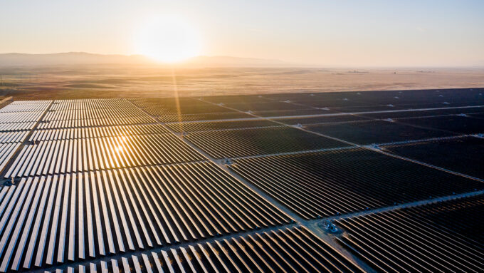 an aerial photo showing a solar farm. There are rows and rows of solar panels grouped into squares as far as the image stretches. The sun is low in the sky.