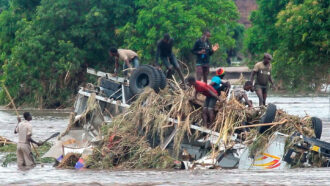 photo of several people standing on an overturned vehicle covered in debris amid floodwaters