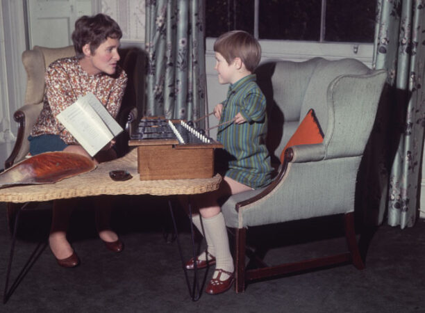 a woman sits and holds up a book to a child with malformed arms playing the xylophone
