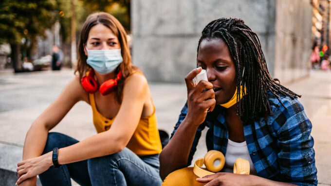 one girl wears a mask seated next to another girl taking an inhaler