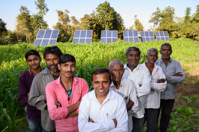 una foto de 8 hombres parados en forma de av frente a un campo con cultivos y paneles solares
