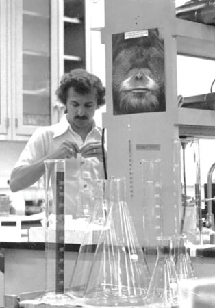 black and white photo of Paul Modrich in a lab in 1977 with a poster of an orangutan in the background