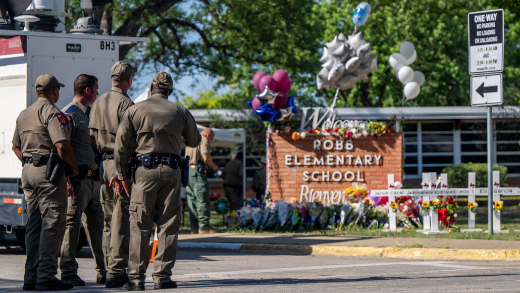 law enforcement officers stand in front of Robb Elementary School where flowers and crosses are displayed in front of a sign reads "Welcome" and "Bienvenidos"