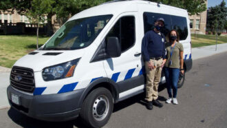 two mental health professionals stand in front of a police van on a street in Denver