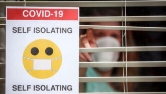 A woman wearing a mask pokes through window blinds. A sign on the window says COVID-19 with SELF ISOLATING above and below a masked face