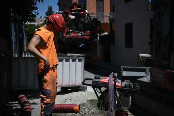 a man dressed in orange and wearing a red helmet wipes sweat from his face at a work site
