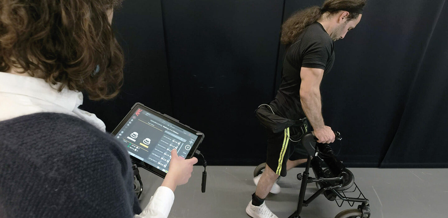 photo of Michel Roccati standing and using a walker as a researcher monitors his progress on an tablet