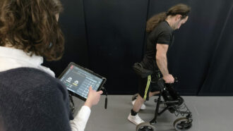 photo of Michel Roccati standing and using a walker as a researcher monitors his progress on an tablet