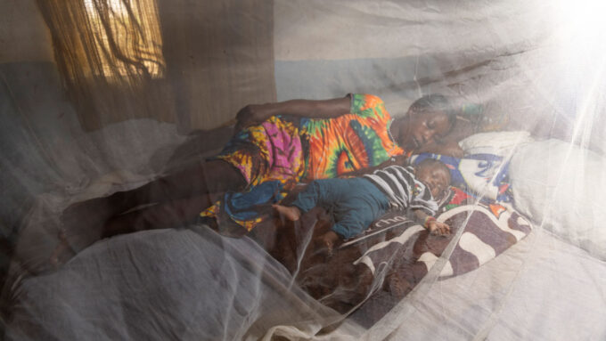 a woman sleeps next to her baby. The photo is taken through a mosquito net.