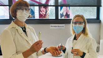 Chemist Michel Nieuwoudt and art historian Erin Griffey, both blond women wearing goggles, masks and lab coats, holding vials and standing in front of Renaissance-era art and a sign that reads Beautiful Chemistry