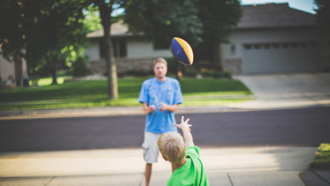 A child throwing a toy football to an older man standing in the driveway of a suburban neighborhood