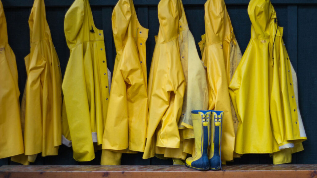 Seven yellow water-resistant jackets hanging up on a rack next to a pair of yellow rain boots