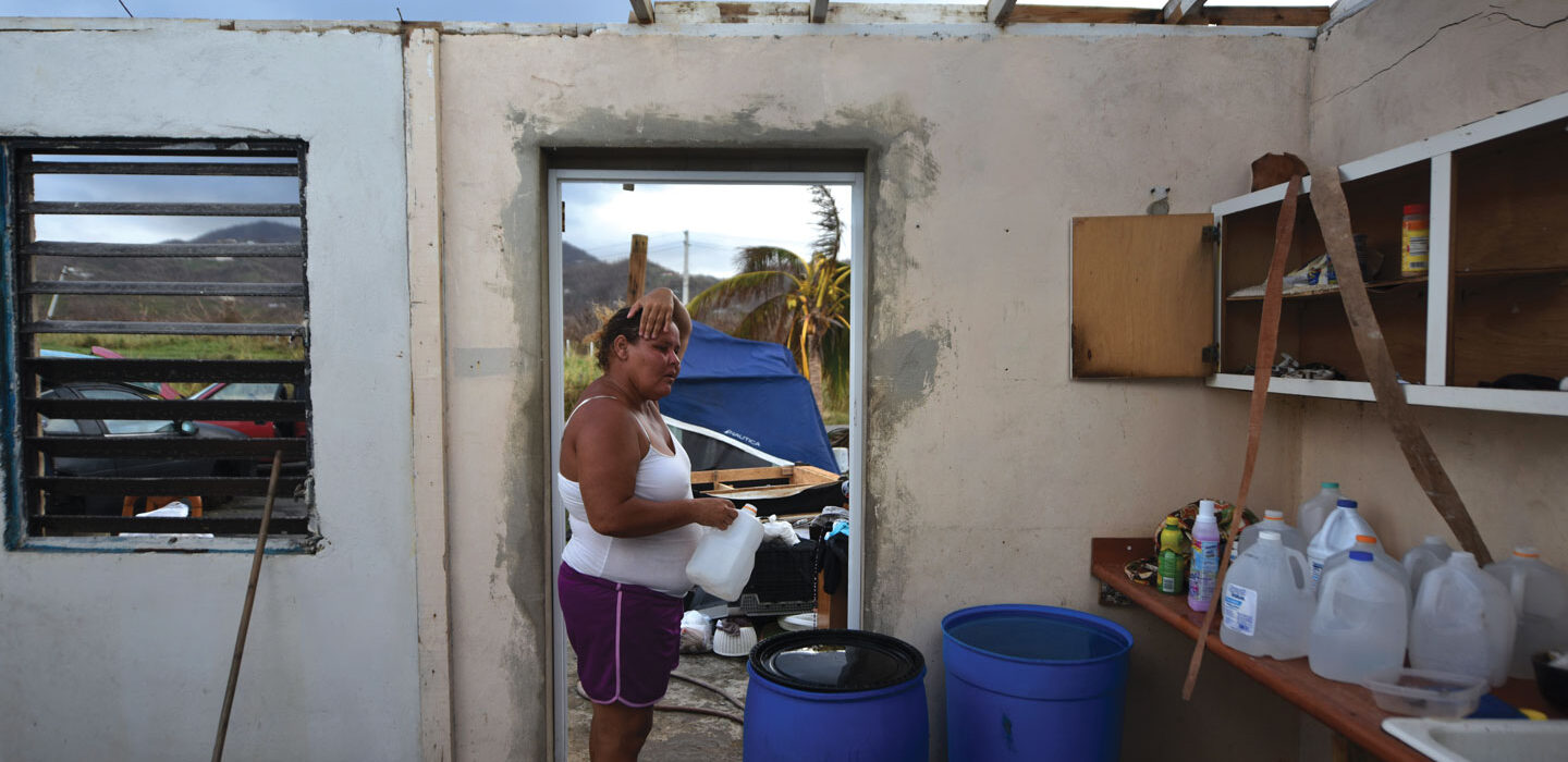 Yasmin Morales stands in the doorway of her home with debris and the Puerto Rico landscape visible behind her. The roof is absent. Jugs of water sit on a counter top, and two big blue barrels stand in front of her.