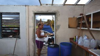 Yasmin Morales stands in the doorway of her home with debris and the Puerto Rico landscape visible behind her. The roof is absent. Jugs of water sit on a counter top, and two big blue barrels stand in front of her.