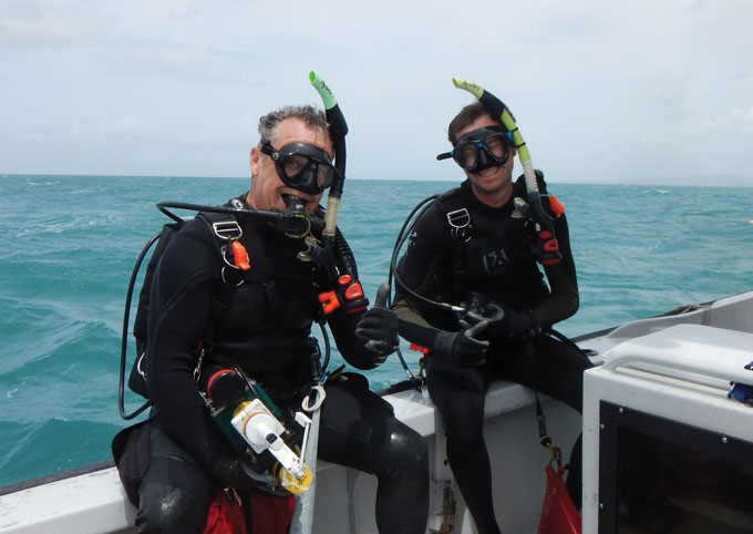 Photo of Curt Storlazzi (left) and Joshua Logan (right) wearing scuba gear and sitting next to each other on the side of a boat in the ocean