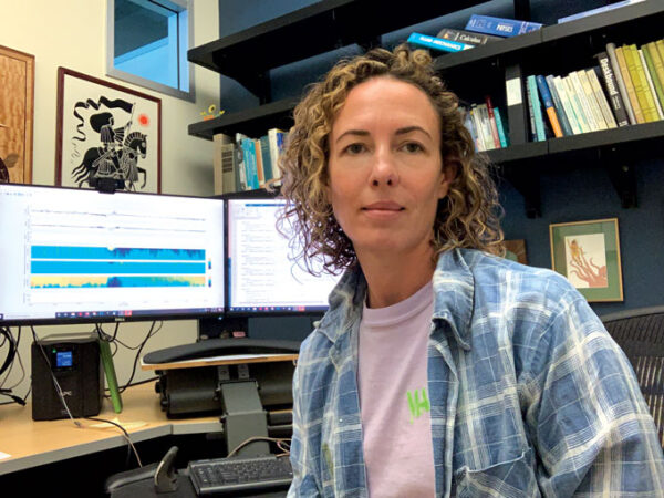 photo of Olivia Cheriton sitting at a desk with two computer monitors and an array of books behind her