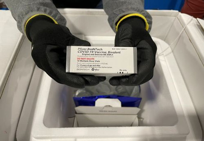 Manos enguantadas sosteniendo una caja que contiene el refuerzo de la vacuna bivalente COVID-19 de Pfizer-BioNTech